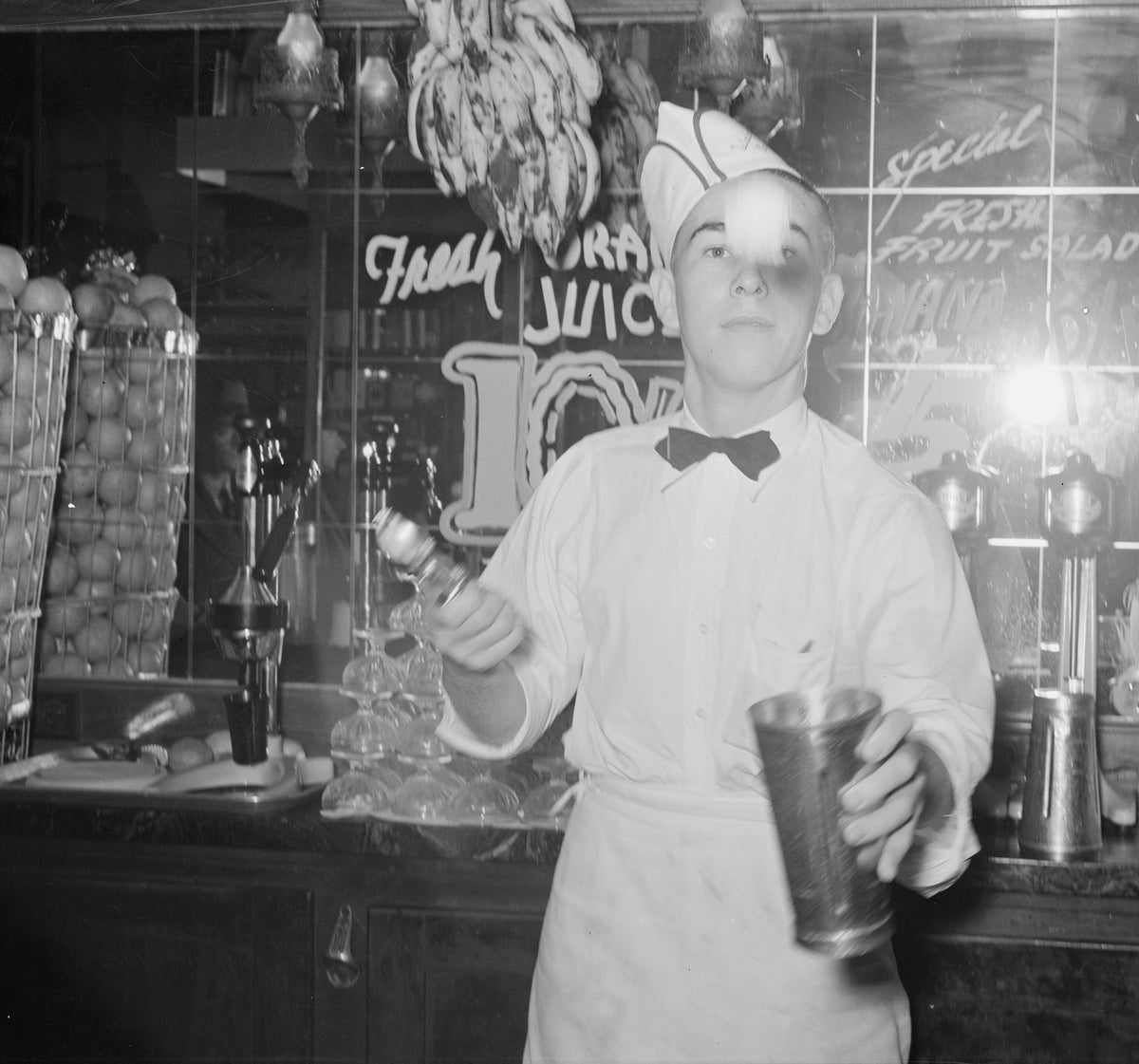 A soda jerk in whites, a paper cap and a bow tie, flipping ice cream into a malted milk shaker behind a drugstore fountain counter. Russell Lee, Corpus Christi, Texas, 1939.